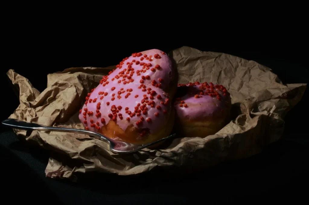 Two heart-shaped donuts with pink frosting and red sprinkles rest on crumpled brown paper. A metal spoon adds a rustic touch to the dark setting.