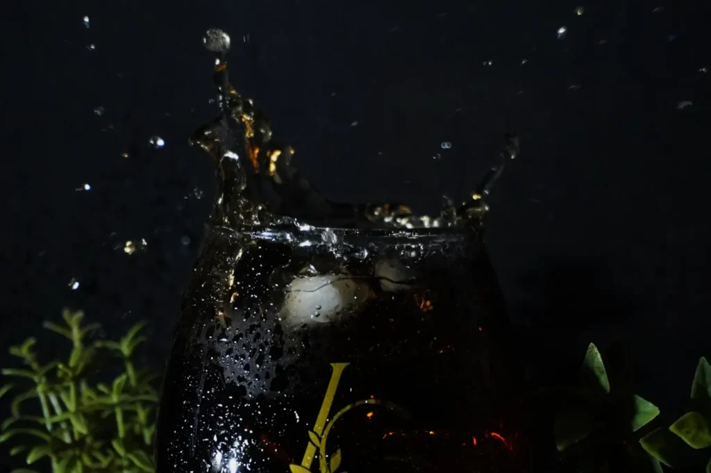 A glass with dark liquid and ice cubes splashes up, droplets frozen mid-air. Set against a dark background with green foliage, creating a dynamic, refreshing scene.