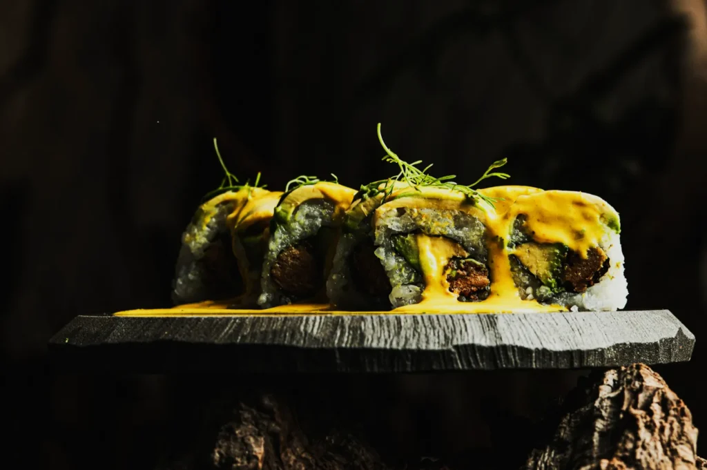 Close-up of sushi rolls on a rustic stone plate, drizzled with vibrant yellow sauce, garnished with fresh herbs, set against a dark, moody background.