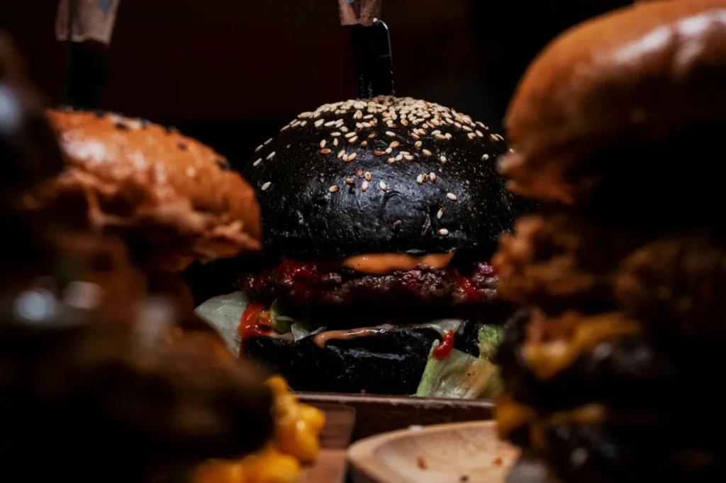 A black sesame-seed burger bun with lettuce, a juicy beef patty, and sauce, accented by a knife, is flanked by two blurred burgers, creating a dramatic, gourmet vibe.