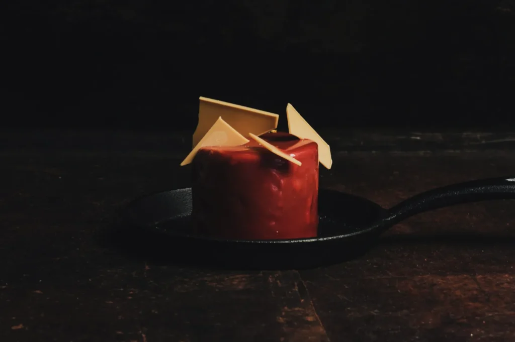 A small, round dessert with a glossy red coating, topped with cream-colored geometric chocolate pieces, sits in a black skillet on a dark wooden table.