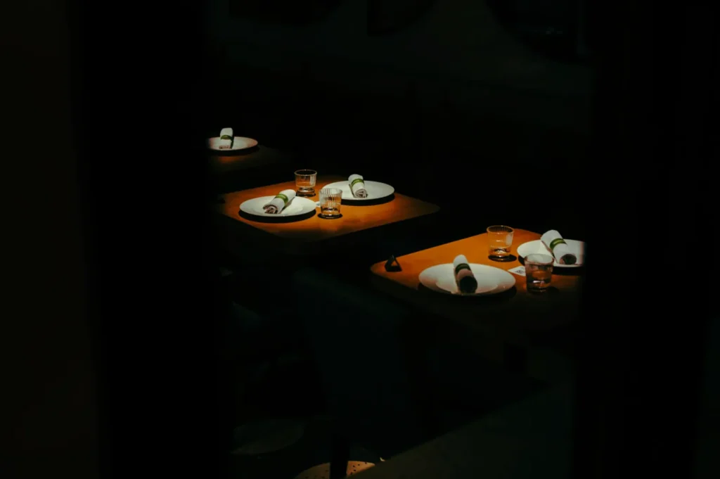 Dimly lit restaurant with neatly set tables, each featuring plates, rolled napkins, and glasses. Mood is cozy and intimate, with focused lighting.