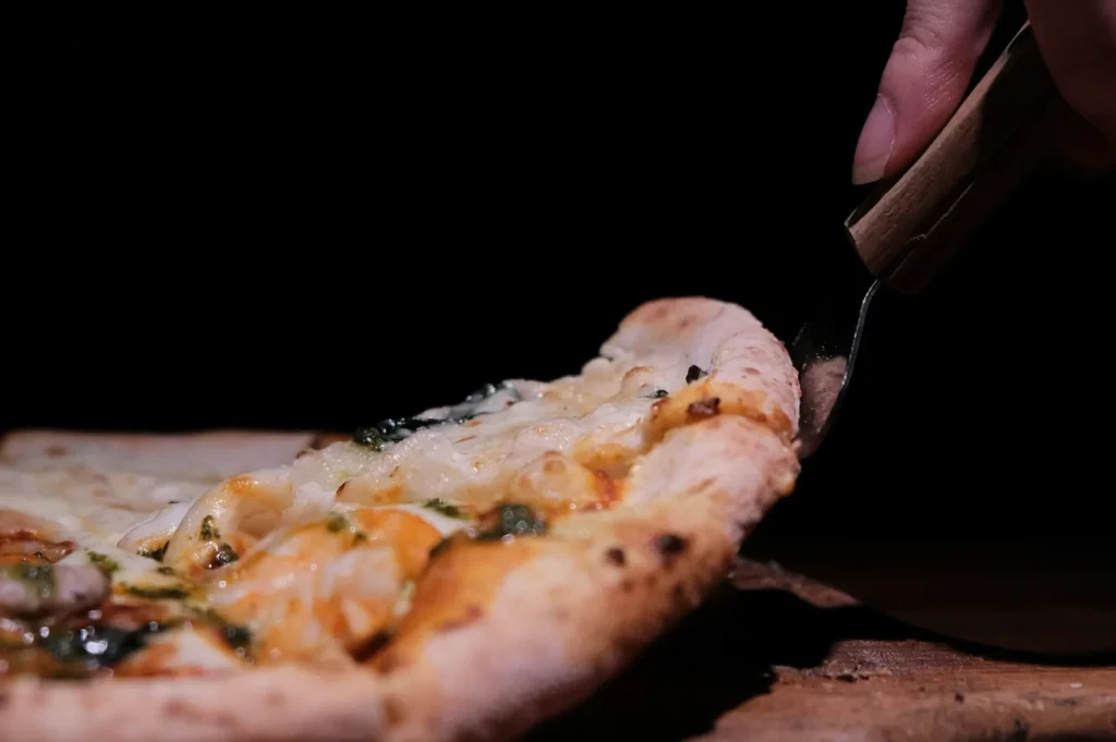 Close-up of a hand slicing a pizza with a knife. The pizza has bubbling cheese and herbs, with a rustic crust, set against a dark background.