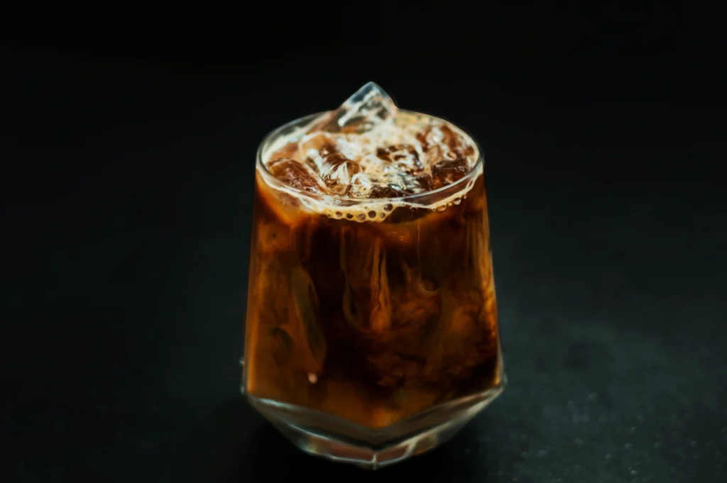 A glass of iced coffee with milk swirls, filled with ice cubes, sits against a dark background. The frothy top adds a refreshing feel.