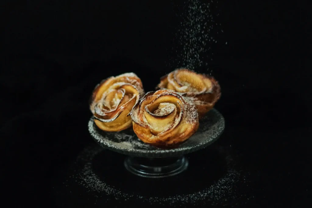 Three apple rose tarts dusted with powdered sugar are elegantly presented on a dark pedestal against a black background, creating a delicate and inviting scene.