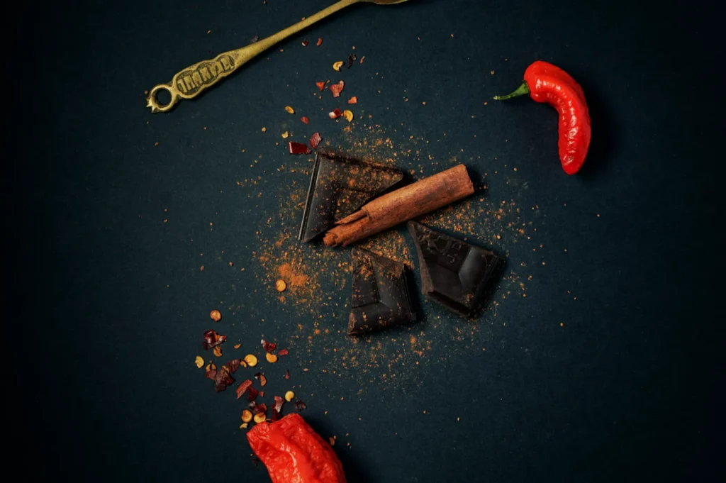 Dark chocolate pieces with a cinnamon stick on a dark background, sprinkled with chili flakes. Two red chili peppers and a vintage spoon add contrast.
