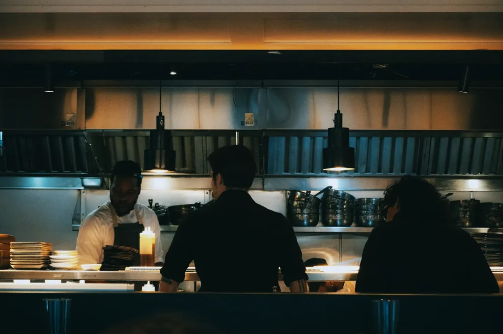 Chef in a sleek restaurant kitchen converses with two patrons. Soft lighting highlights stacked plates and hanging utensils, creating a warm, bustling atmosphere.