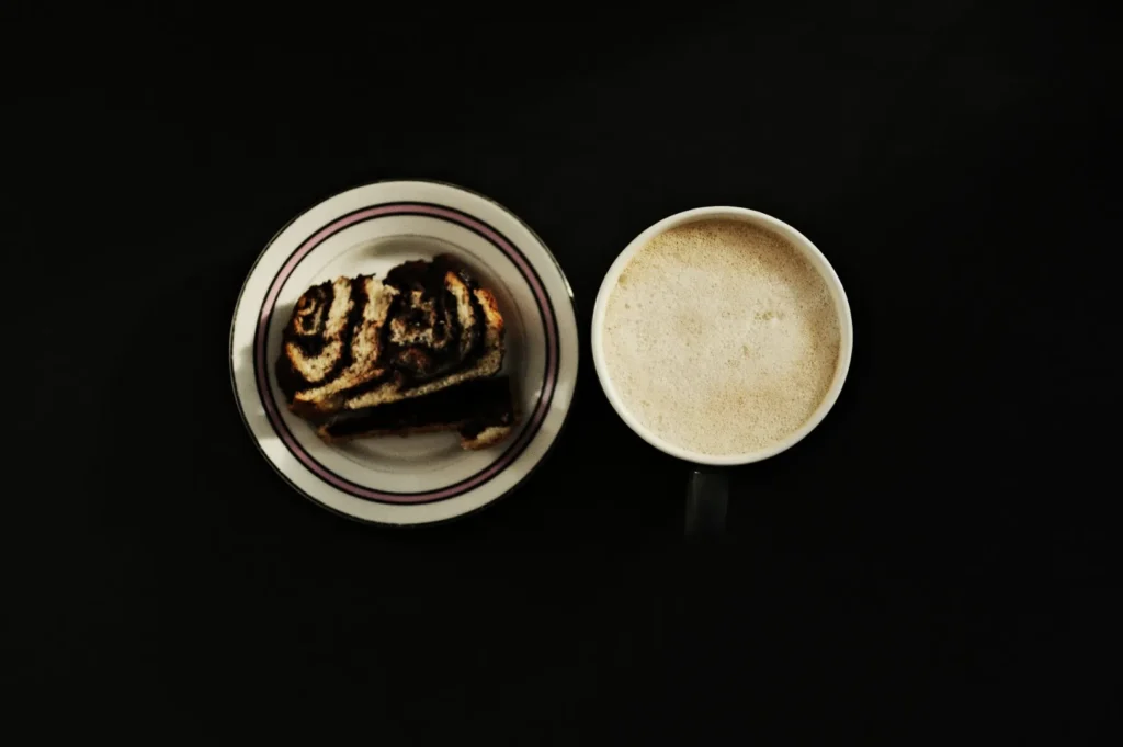 A cup of frothy coffee sits next to a plate with two slices of swirl-patterned bread on a dark background, creating a cozy breakfast scene.