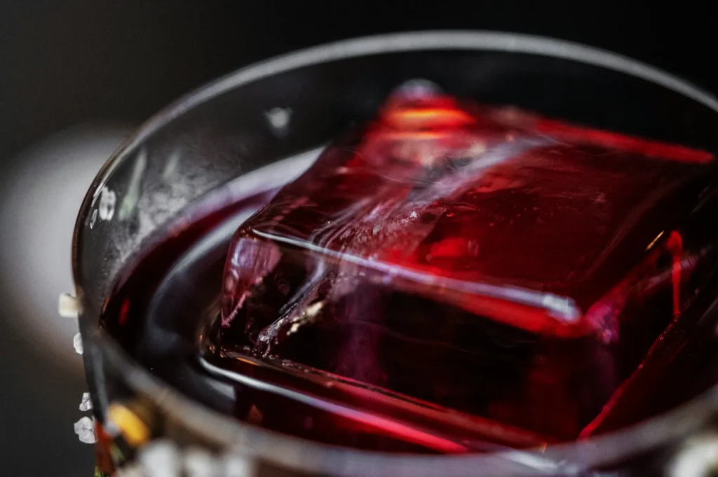 Close-up of a clear glass with a large red ice cube submerged in a dark liquid, possibly a cocktail. The image conveys a sophisticated and refreshing tone.