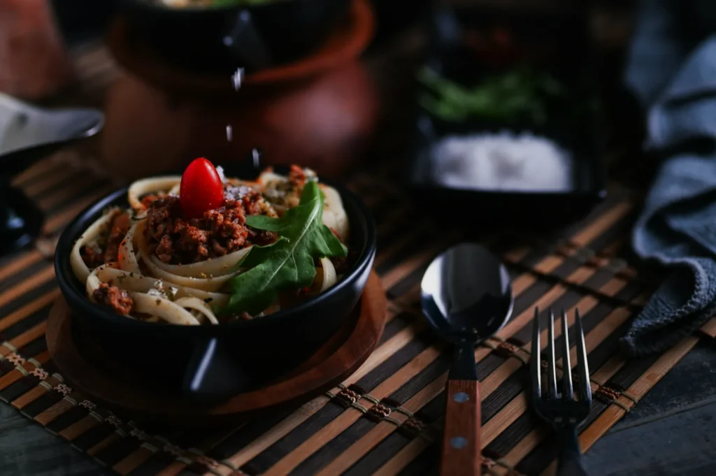 A bowl of pasta with meat sauce, topped with a cherry tomato and arugula, sits on a bamboo mat. Salt is being sprinkled above. A fork and spoon rest nearby, creating a cozy dining atmosphere.