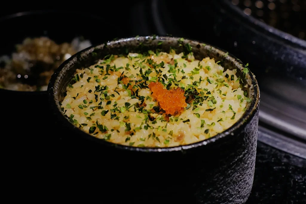 A textured black bowl filled with creamy risotto, garnished with chopped green herbs and a small mound of orange fish roe on top, against a dark background.