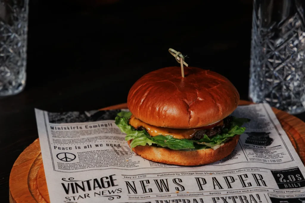 A cheeseburger with lettuce and a wooden skewer on a mock vintage newspaper-lined wooden plate. Two crystal glasses are partially visible.