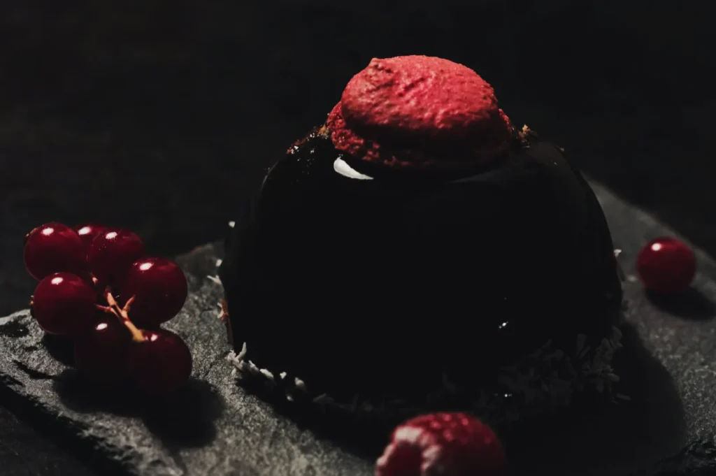 A glossy, dark chocolate dessert dome topped with a red macaron sits on a slate plate, surrounded by red currants and raspberries, creating a rich, elegant tone.