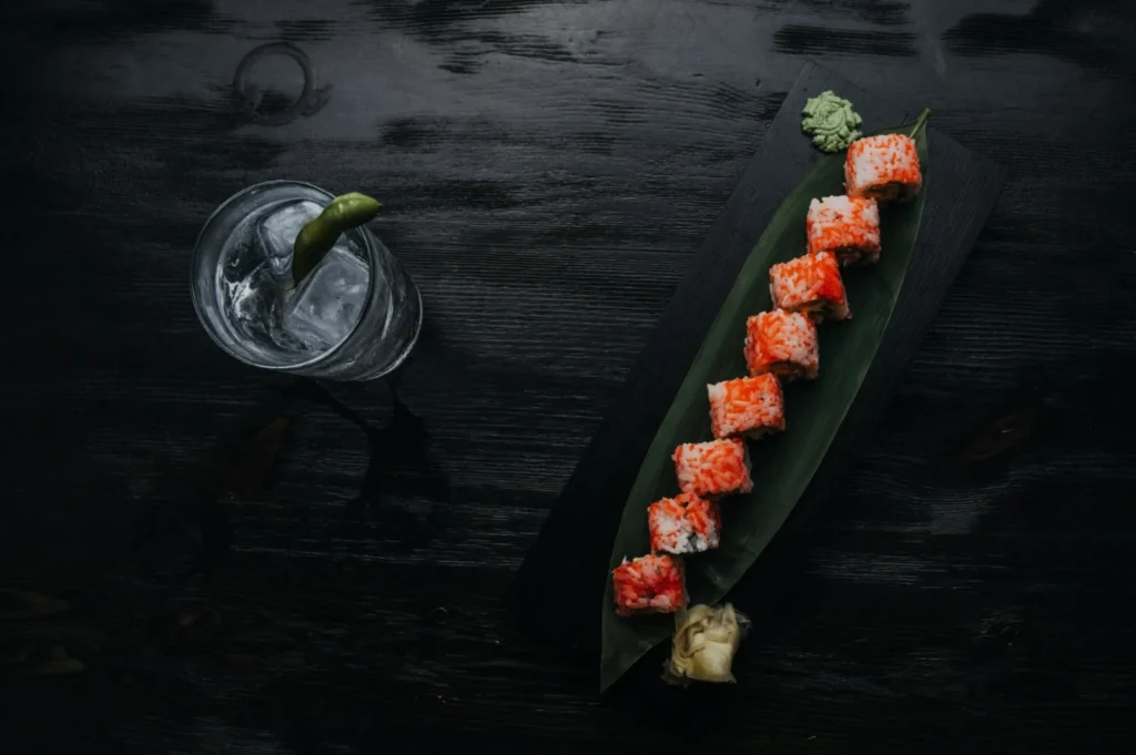 Sushi rolls with red toppings on a dark platter with green leaf, garnished with wasabi and ginger, beside a drink with ice and a lime wedge.