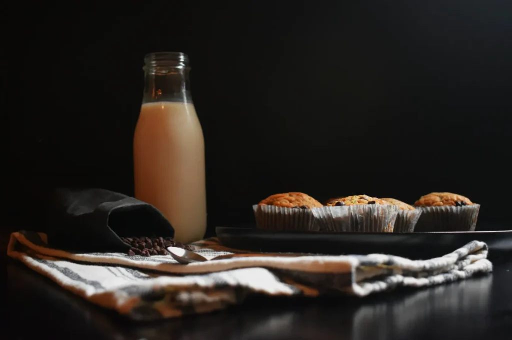 A bottle of milk and chocolate chip muffins on a dark plate sit on a striped cloth. Nearby, an open black pouch with chocolate chips spills out, creating a cozy, warm atmosphere.
