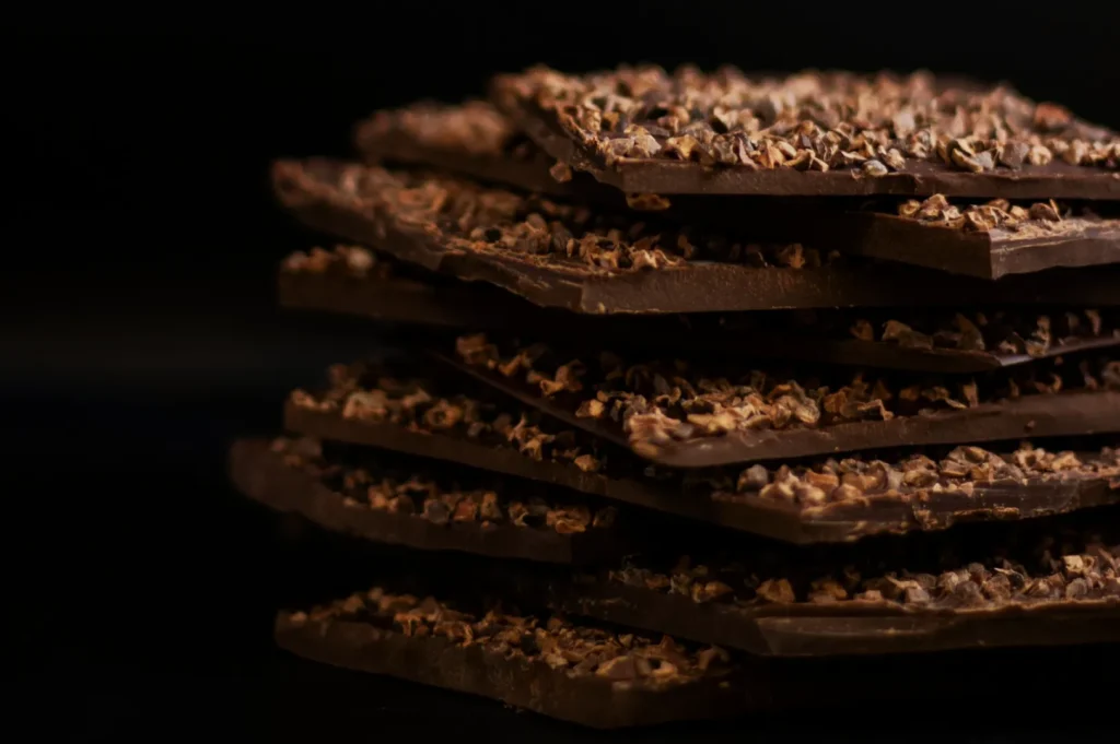 Stack of dark chocolate bark pieces, topped with crushed nuts, on a dark background. The rich texture and indulgent tone evoke luxury.