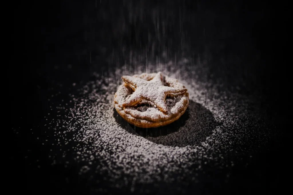 A single star-topped mince pie is dusted with powdered sugar, set against a dark background, creating a cozy and festive atmosphere.