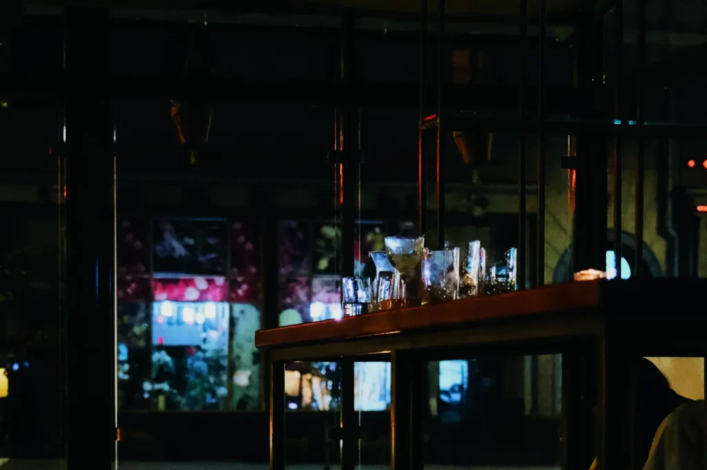 Dimly lit bar interior with scattered empty glasses on a high shelf. Outside, blurred colorful lights and wall art create a moody, quiet ambiance.