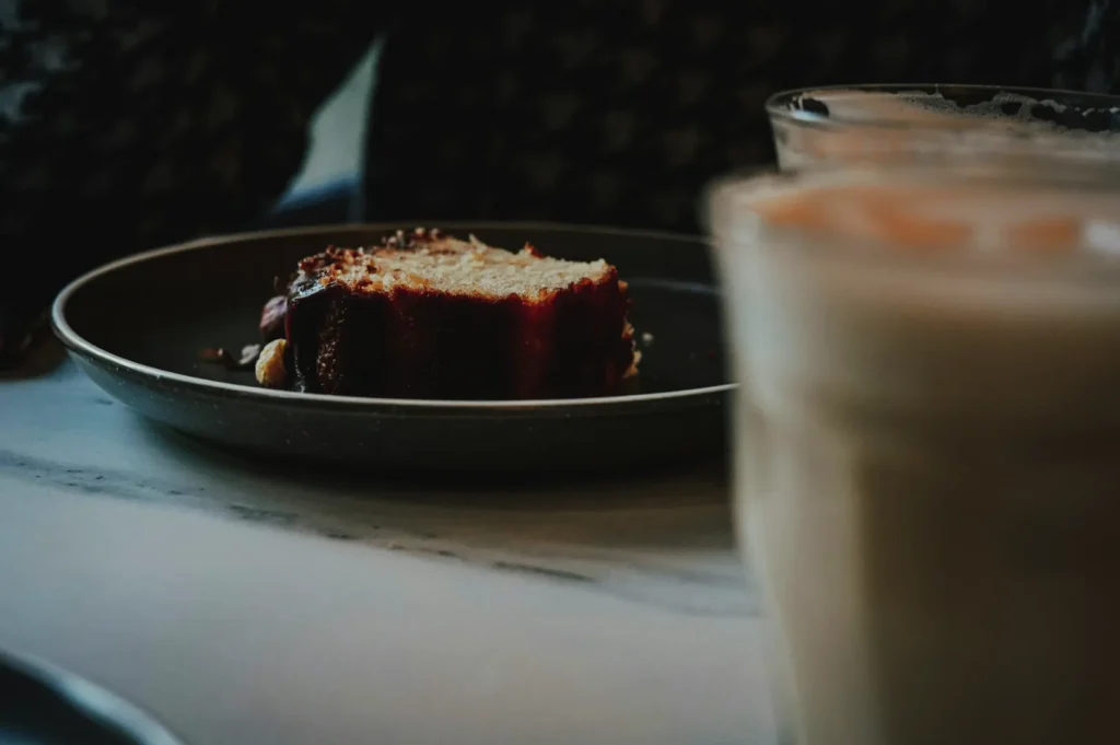 A slice of cake with dark glaze sits on a dark plate, with two blurred glasses in the foreground. The lighting is dim, creating a cozy atmosphere.