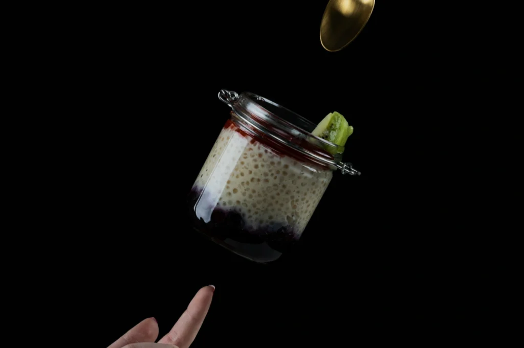 A jar of chia pudding with a fruit topping, including kiwi and a red sauce, is suspended mid-air, with a hand pointing upwards and a gold spoon above.