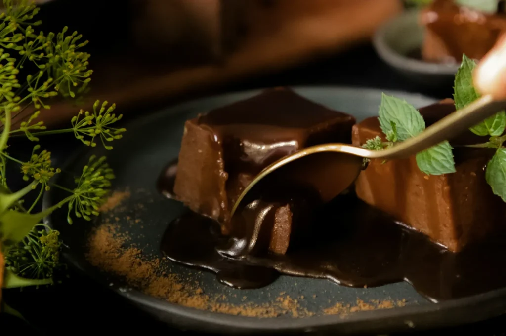 A spoon cuts into rich chocolate cake squares drizzled with glossy sauce on a dark plate, garnished with fresh mint leaves and cinnamon, creating an indulgent, elegant dessert scene.
