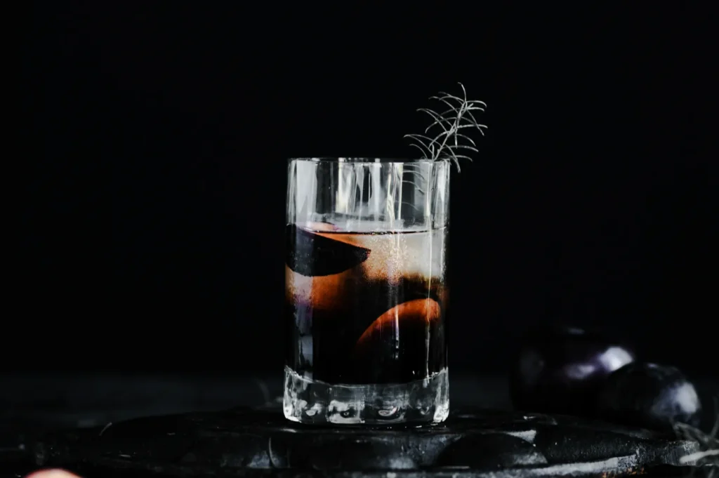 A clear glass filled with dark liquid and plum slices is garnished with a sprig against a black background, creating a moody, sophisticated ambiance.
