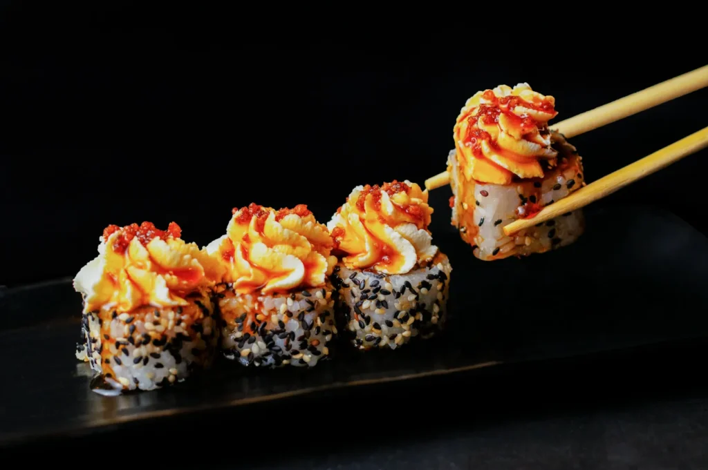 Sushi rolls on a black platter, topped with creamy orange sauce and red garnish, with chopsticks holding one piece. Elegant, appetizing presentation.