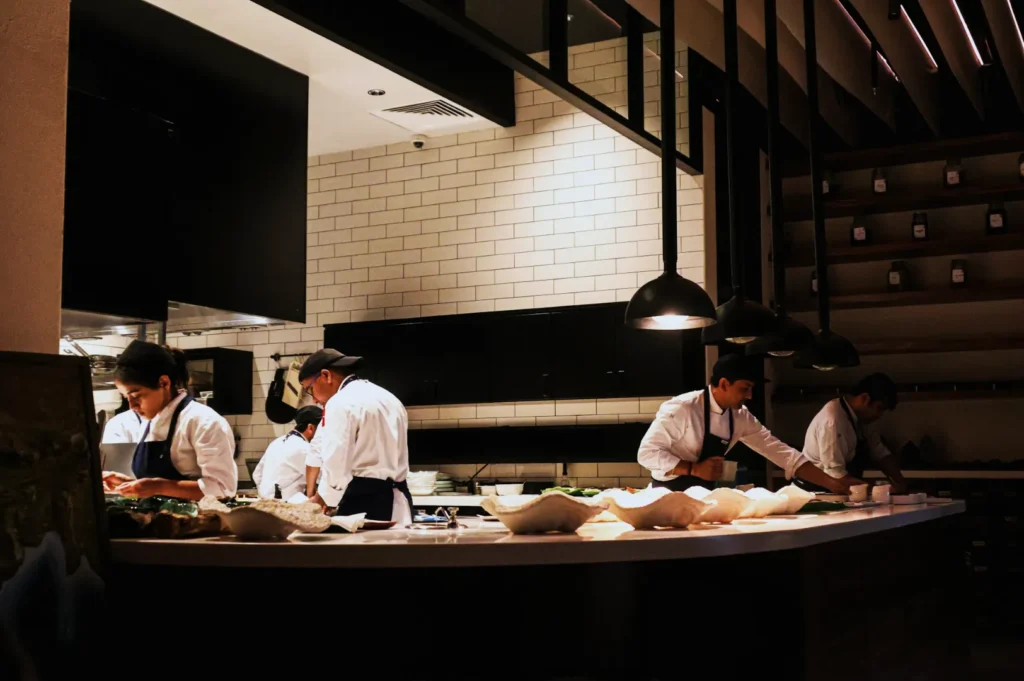 Chefs in white uniforms and dark aprons prepare food in a modern kitchen with subway tiles, dim lighting, and organized shelves, exuding a focused ambiance.