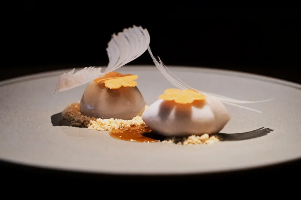 Elegant dessert presentation on a gray plate featuring two dome-shaped confections topped with delicate feathers and flower-shaped crisps, set on a bed of crumbs.