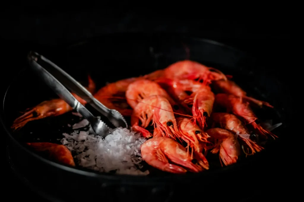 Cooked shrimps arranged in a black pan with coarse sea salt and metal tongs. The scene is set against a dark background, creating a rustic feel.