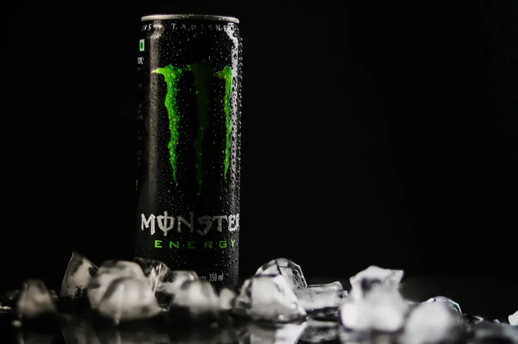 A chilled can of Monster Energy stands on a surface scattered with ice cubes. The can's black and green design contrasts against a dark background.