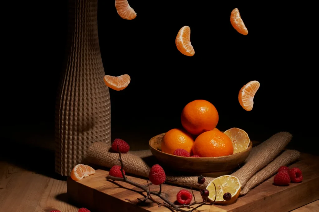 A still life image featuring a wooden bowl of oranges on a table, surrounded by raspberries and a halved lemon. Tangerine slices appear to float above, set against a dark background, creating a dynamic yet serene atmosphere.