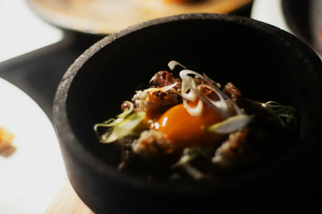 A black bowl filled with a dish featuring a raw egg yolk, sliced green onions, grilled meat, and garnish. The setting is dimly lit, creating a cozy, intimate atmosphere.