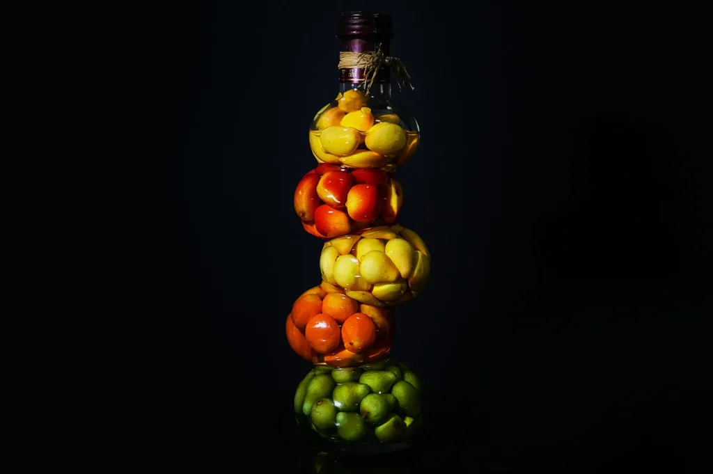 A tall, decorative bottle filled with layered green, orange, and yellow fruits, set against a dark background, conveying a vibrant, artistic display.