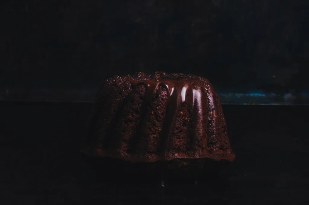 Dark chocolate bundt cake with glossy chocolate glaze, set against a moody, dark background. The rich texture conveys indulgence and warmth.