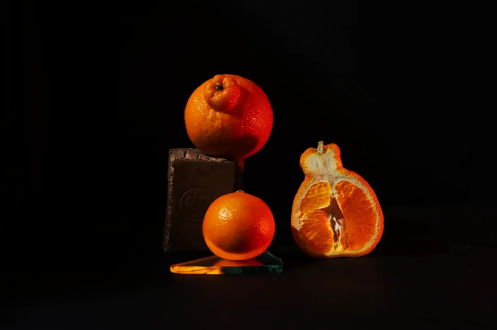 Two oranges and a half-orange are artfully arranged with a chocolate block on colorful glass, under dramatic lighting against a dark background.
