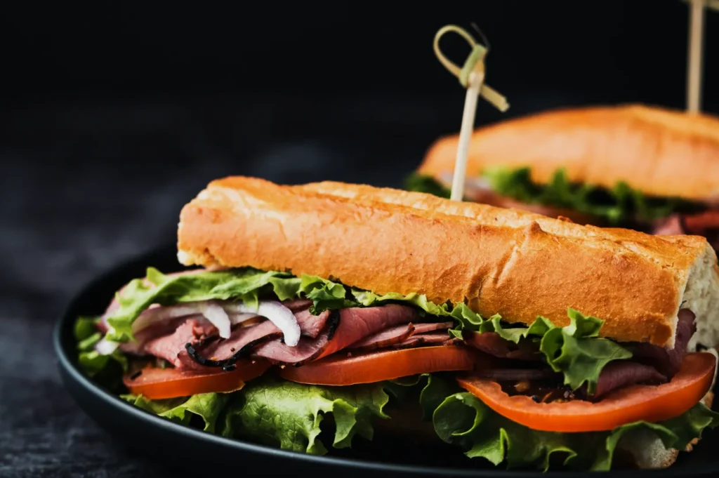 A close-up of a fresh sub sandwich on a black plate. It features roast beef, lettuce, tomato, and onions between a crispy baguette, secured with a wooden skewer.