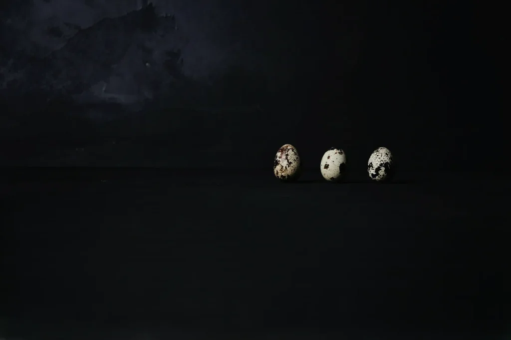 Three speckled quail eggs are placed in a row against a dark, textured background. The lighting casts a dramatic, minimalist tone over the scene.