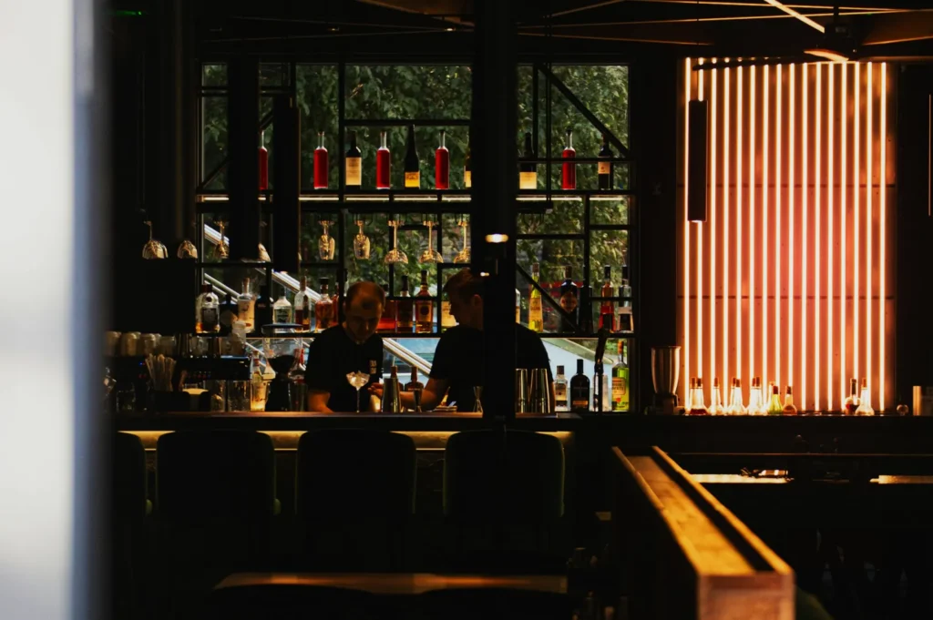 Dimly lit bar with two bartenders at work, surrounded by various bottles on shelves. Warm lighting and tall windows create a cozy, intimate ambiance.