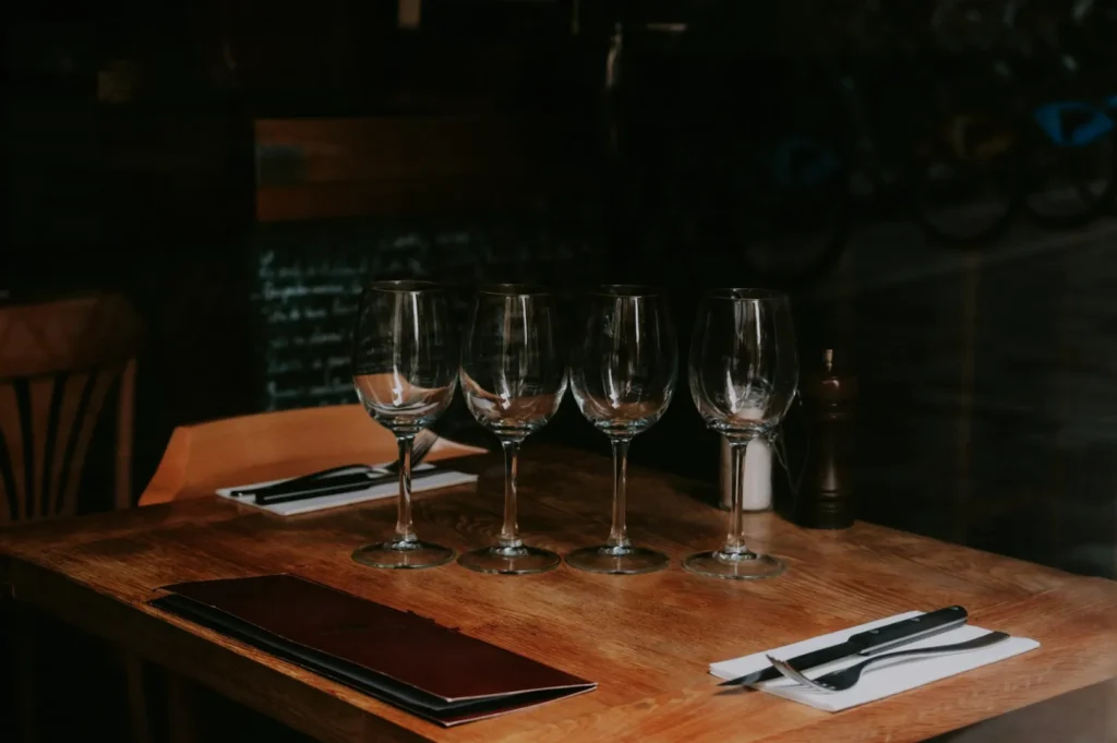 A warmly lit wooden table set for four in a cozy restaurant, featuring empty wine glasses, napkins with cutlery, a menu, and a pepper grinder.