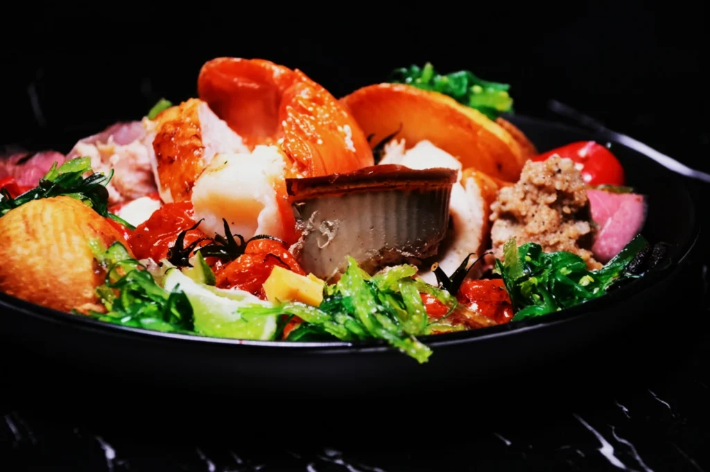 A vibrant dish features assorted seafood including lobster, surrounded by colorful greens, cherry tomatoes, and herbs on a dark plate on a marble surface.