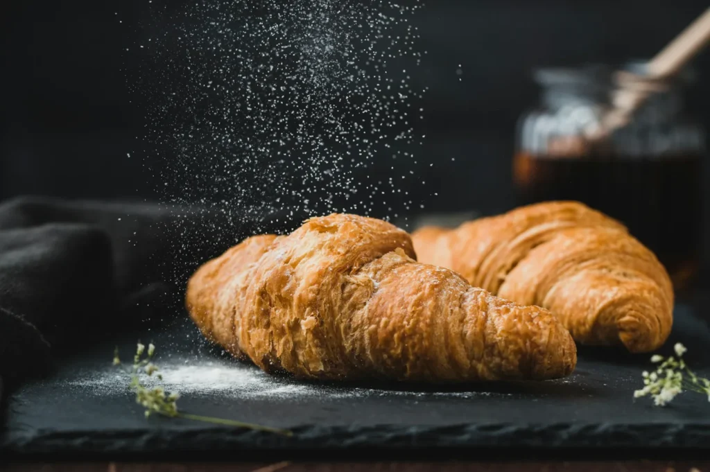 Flaky croissants on slate with sugar dusting, set against a dark background. A honey jar and a black napkin add a rustic, cozy feel.