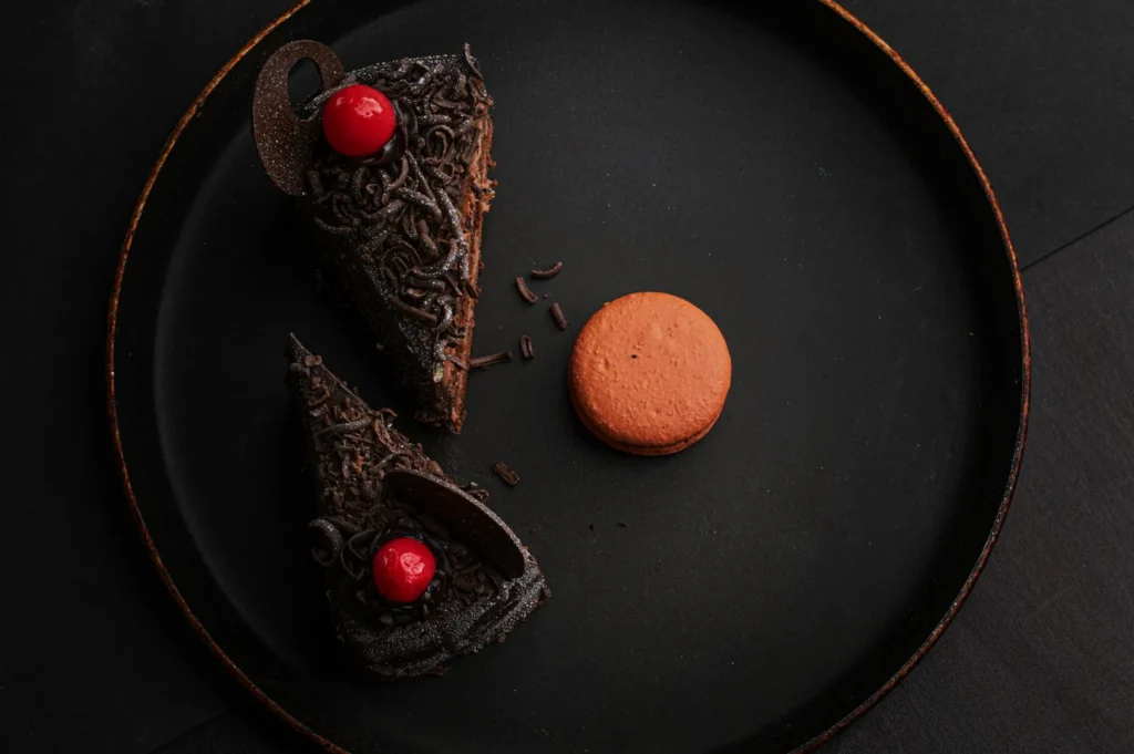 Two slices of rich chocolate cake with cherries and sprinkles on a dark plate, paired with an orange macaron, creating a moody, indulgent vibe.