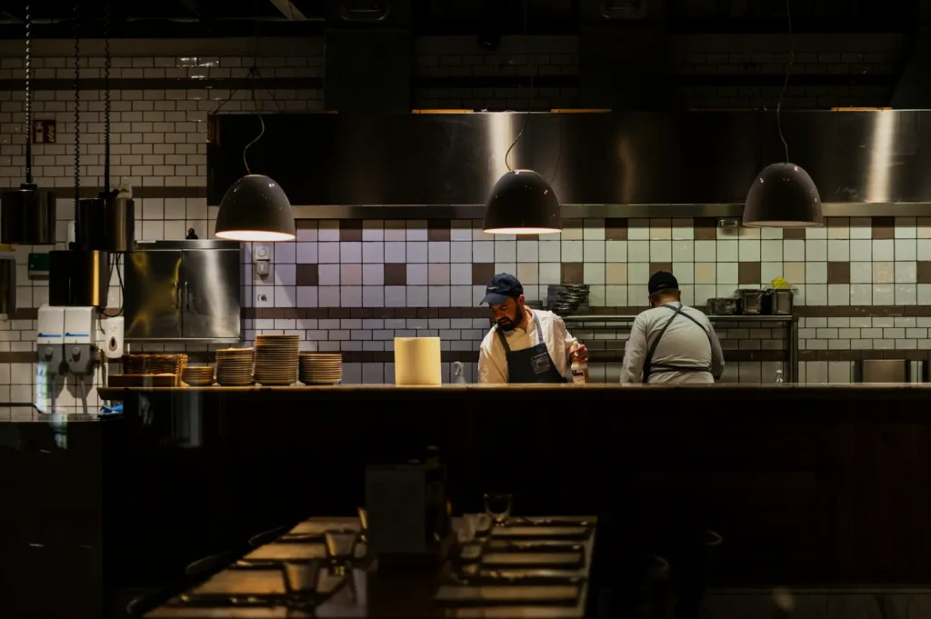 Chefs in a dimly lit kitchen work under large pendant lights, surrounded by white tiles and stacked plates, creating a focused, industrial atmosphere.