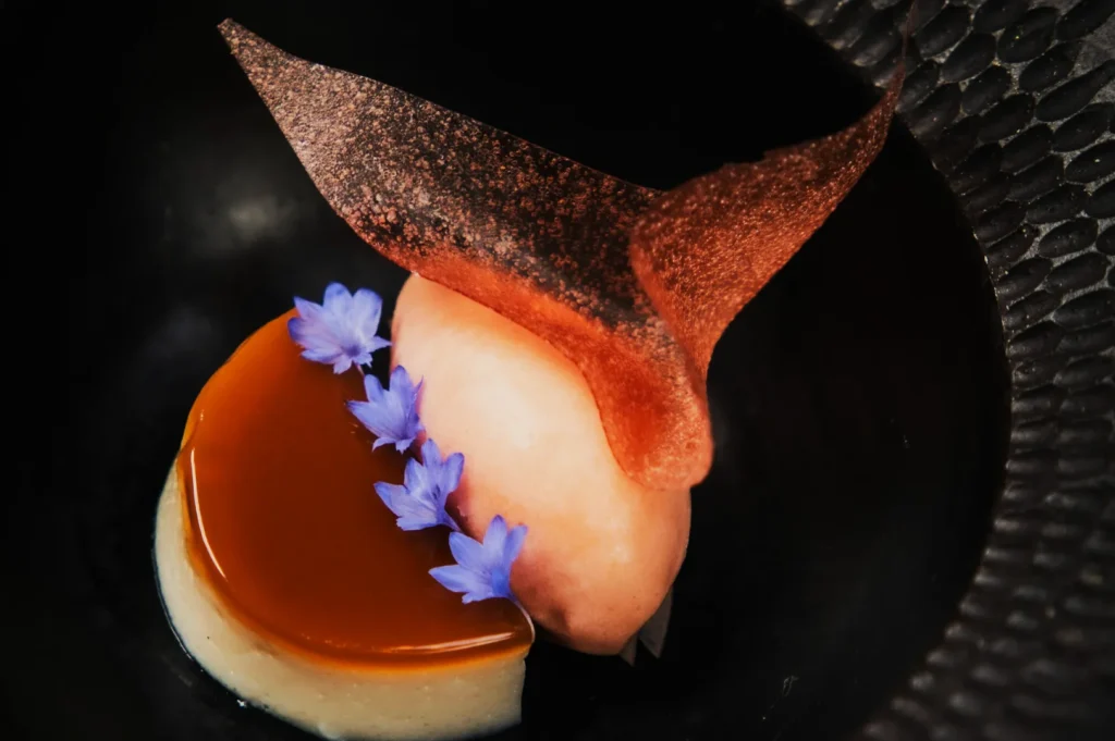 A delicate dessert features a caramel panna cotta, peach sorbet, and a pink sugar shard, garnished with purple edible flowers on a textured black plate.
