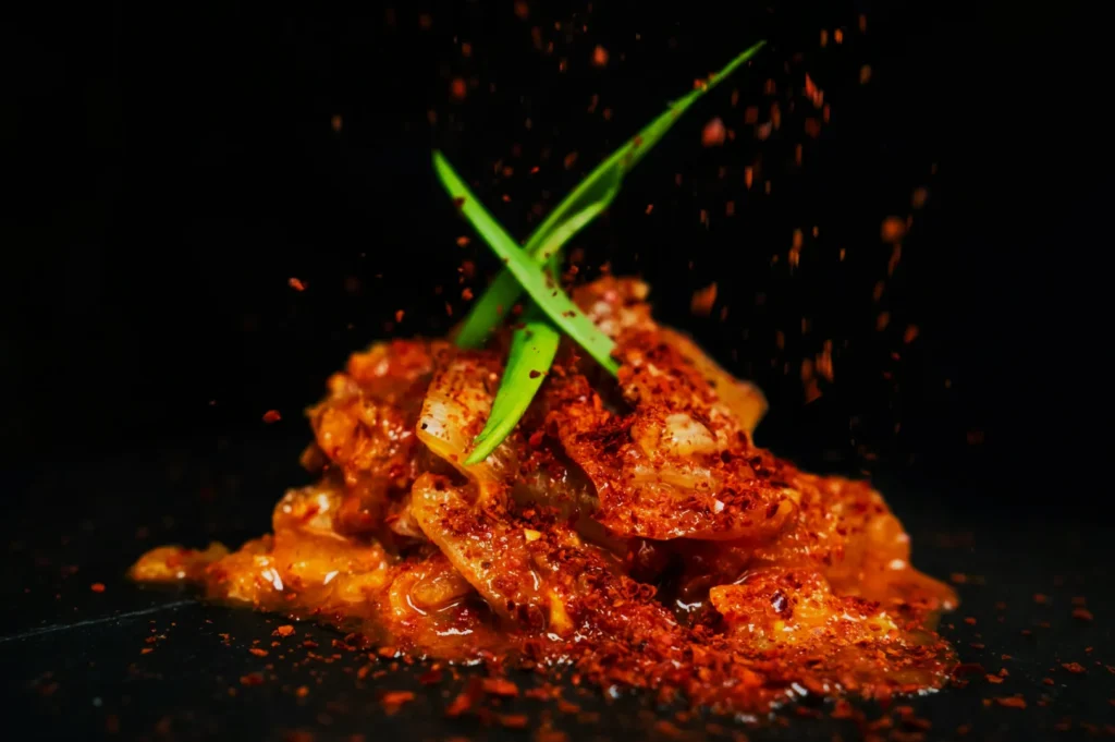 Spicy marinated chicken on a dark plate with vibrant chili flakes scattered above. Two green herbs are crossed on top. The dish looks flavorful and dynamic.