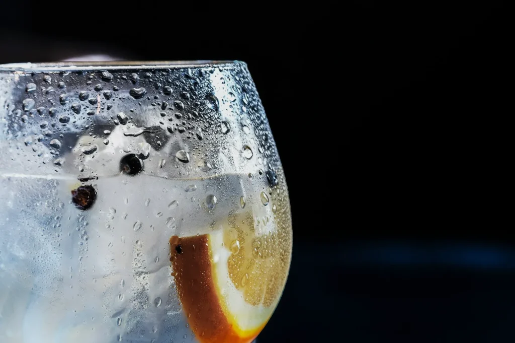 A close-up of a chilled glass with condensation, containing a lemon slice and ice cubes. The refreshing drink conveys a cool, crisp feel against a dark background.