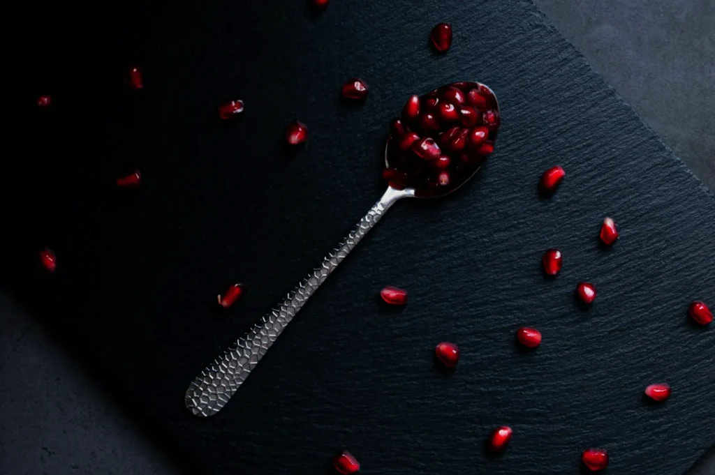 A textured silver spoon filled with glossy red pomegranate seeds on a dark slate surface, surrounded by scattered seeds, conveying elegance and simplicity.