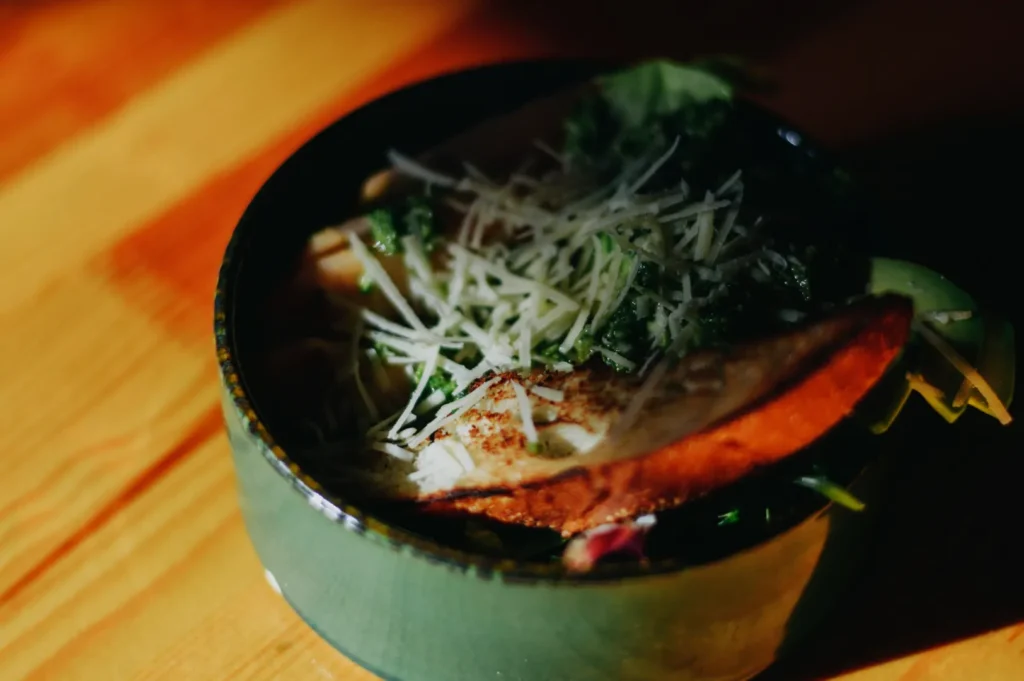 A green bowl with a vibrant salad topped with grated cheese, toasted bread, and a drizzle of green dressing, set on a wooden table with warm lighting.