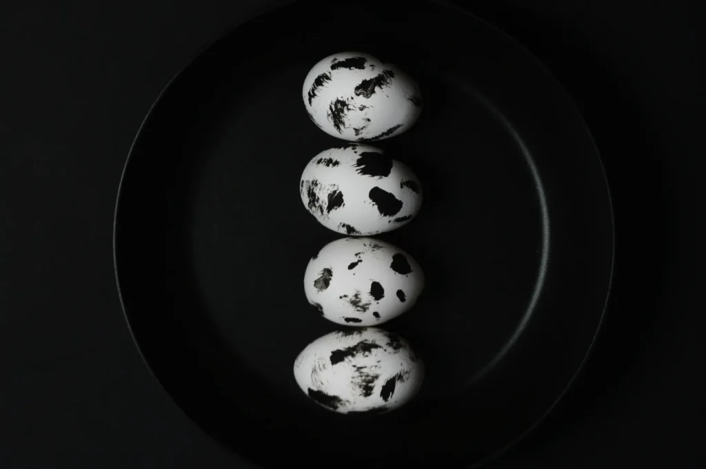 Four white eggs with black speckles are arranged in a row inside a dark frying pan against a black background, creating a minimalist, artistic look.